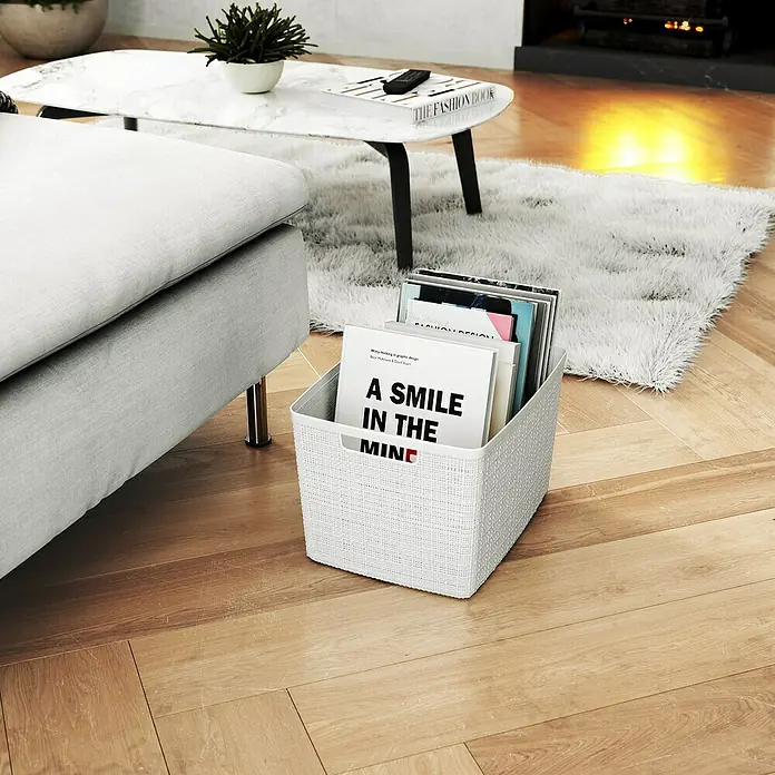 Curver Aufbewahrungsbox Jute - L x B x H: 36 x 28 x 23 cm, Kunststoff, Creme - Ein weißer Korb mit dem Aufdruck "A Smile in the Mind" steht neben einem Sofa, gefüllt mit Büchern, auf einem Holzboden.