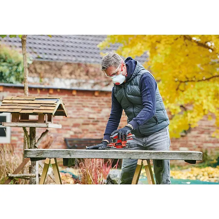 Einhell PXC 18V Lijadora orbital de batería TC-OS 18/187 Li Solo - 18 V, Sin batería, Número de oscilaciones marcha en vacío: 24.000 r.p.m. - Un hombre lija madera con una lijadora eléctrica roja Einhell al aire libre, con equipo de seguridad. Vegetación otoñal al fondo.