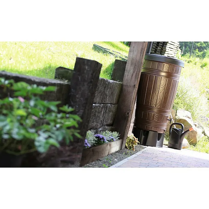 Regenspeicher Woodcan - 265 l, Braun - Eine Regentonne in Holzoptik steht neben einer Mauer und einem schwarzen Gießkanne. Ideal zur Gartenbewässerung und Regenwassernutzung.