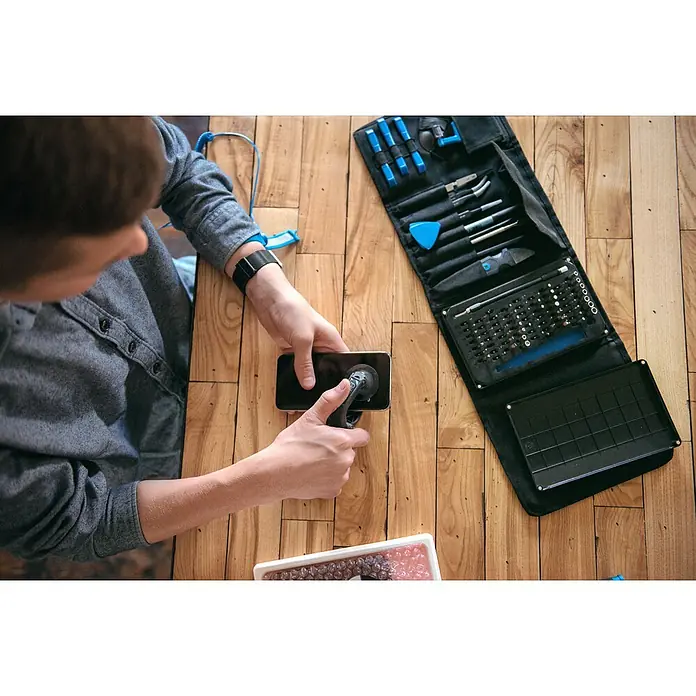 iFixit Set de herramientas Pro Tech - 84 pzs. - Reparación de un smartphone con herramientas sobre una mesa de madera, con un kit de herramientas y pulsera de conexión a tierra.