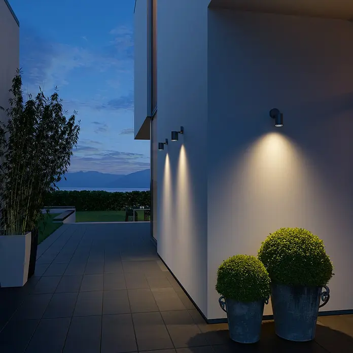 Steinel Aplique exterior LED con sensor Spot One S - 7,47 W, L x An x Al: 9,8 x 9,7 x 17,5 cm, Antracita, Blanco cálido - Exterior de una casa moderna con luces de pared exteriores que iluminan la fachada, flanqueada por vegetación.