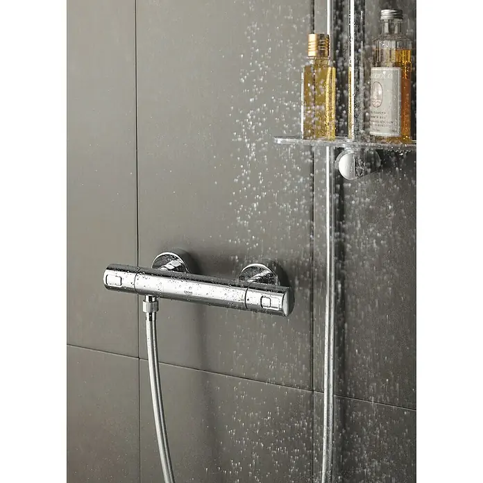 Grohe Thermostatarmatur Precision Joy - Breite: 29,9 cm, Chrom, Glänzend - Nahaufnahme eines verchromten Duschthermostats mit Wasser, daneben Shampoo-Flaschen auf einem Regal in einer modernen Duschkabine.