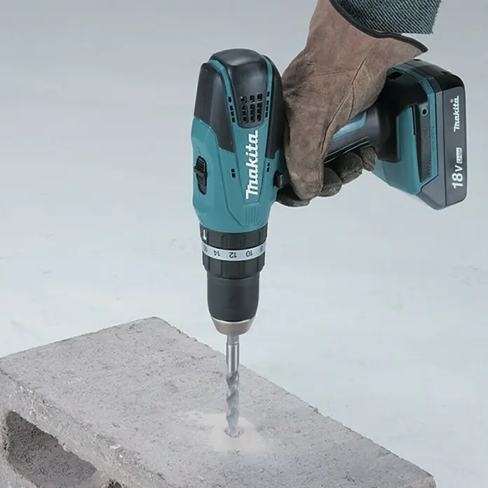 Makita Taladro atornillador percutor de batería HP457DWE - 18 V, 2 baterías, 1,5 Ah, 42 Nm - Un taladro inalámbrico Makita en acción, perforando un agujero en un bloque de hormigón. Perfecto para construcción y bricolaje.