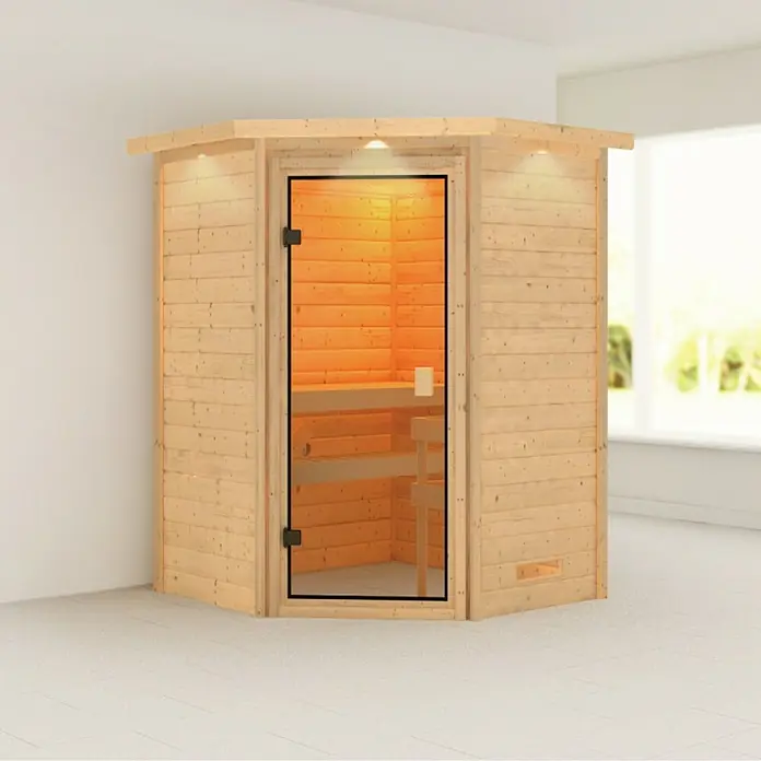 Karibu Massivholzsauna Antonia - Ohne Ofen, L x B x H: 159 x 173 x 191 cm, Mit Dachkranz und Beleuchtung - Sauna-Ecke aus hellem Holz mit Glastür in einem hellen Raum. Die Sauna ist mit Bänken und Beleuchtung ausgestattet.