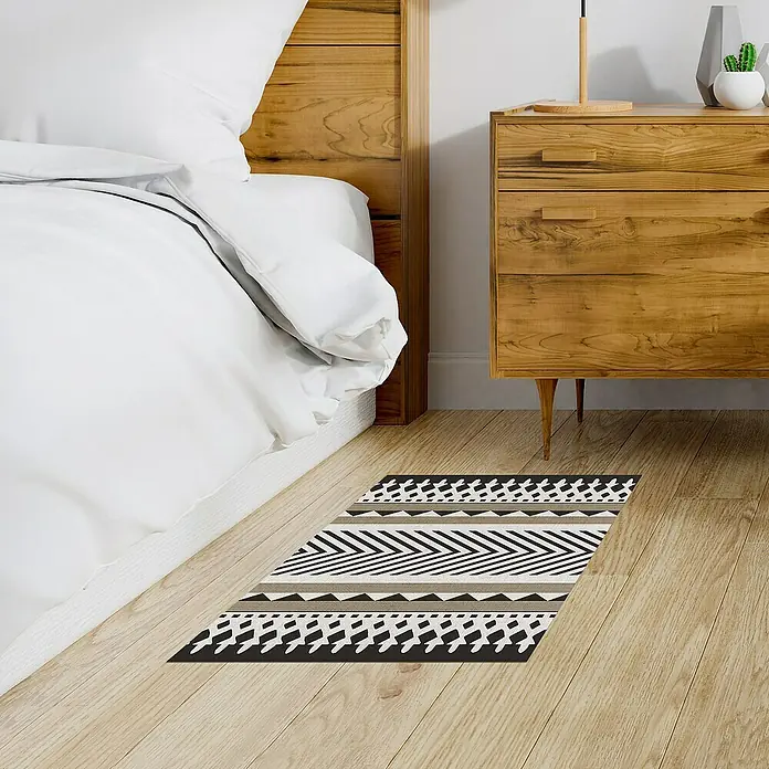 Alfombra de vinilo Croma Etnic stone - Blanco/Marrón/Negro, 75 x 45 cm - Vista de una alfombra estampada frente a una cama y una cómoda de madera. La alfombra presenta un diseño moderno y llamativo.