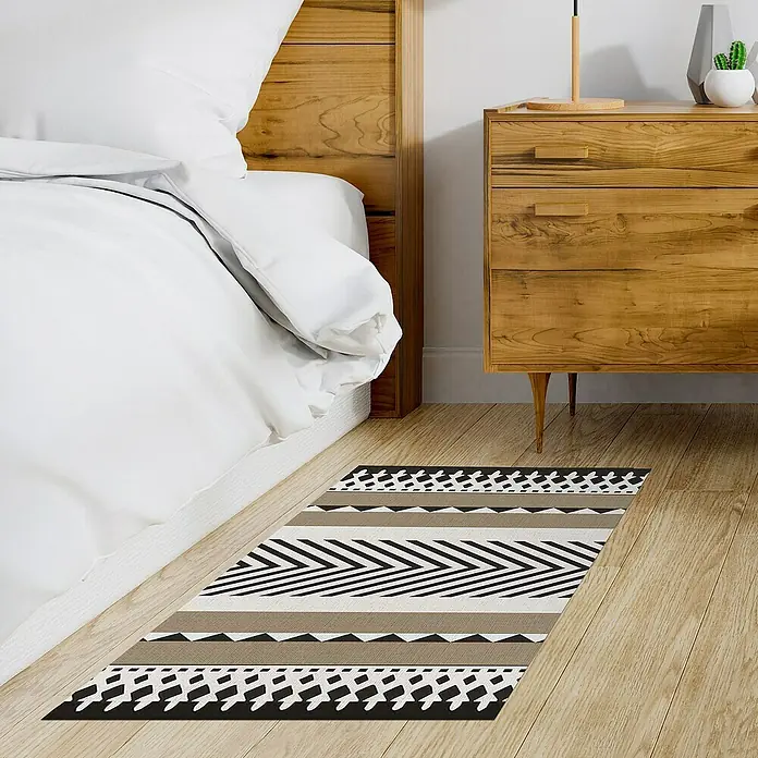 Alfombra de vinilo Croma Etnic Stone - Blanco/Marrón/Negro, 110 x 50 cm - Una alfombra decorativa con un patrón geométrico se encuentra junto a una cama y una cómoda sobre un piso de madera. Es elegante y llamativa.