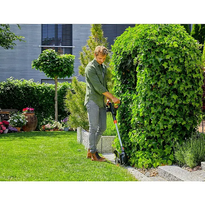 Gardena Power for All 18V Akku-Rasentrimmer ComfortCut - 18 V, Li-Ionen, Ohne Akku, Schnittbreite Messer: 23 cm - Ein Mann trimmt mit einer Akku-Grasschere präzise die Rasenkante unter einem grünen Busch in einem gepflegten Garten.