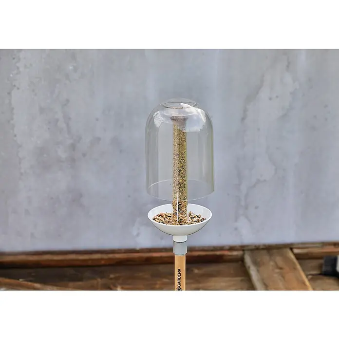 Gardena ClickUp! Vogelfutterstation - 15,7 x 15,7 x 35,2 cm, Kunststoff - Nahaufnahme einer Gardena Vogelfutterstation mit durchsichtigem Dom und gefülltem Futterrohr, die auf einem Holzstab im Garten steht.