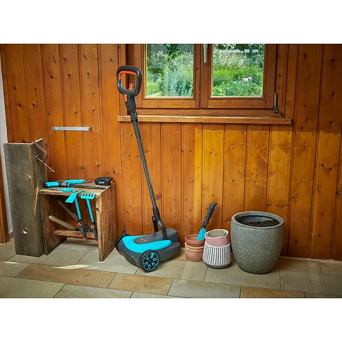 Gardena Power for All 18V Akku-Rasenmäher - 18 V, Ohne Akku, 2,5 Ah, Schnittbreite: 22 cm - Eine Hofkehrmaschine von GARDENA steht zusammen mit Gartengeräten und Blumentöpfen vor einer Holzwand in einem Gartenhaus.