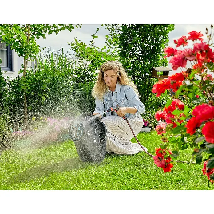 Gardena Mähroboter Sileno minimo 500 - 500 m², Schnittbreite: 16 cm, Induktionsgestützte Navigation, Begrenzungskabel - Eine Frau reinigt eine GARDENA-Schlauchtrommel mit einem Wasserschlauch im Garten, umgeben von üppigem Grün und Blumen.