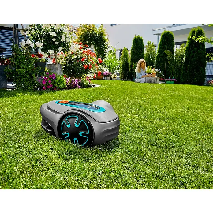 Gardena Mähroboter Sileno minimo 500 - 500 m², Schnittbreite: 16 cm, Induktionsgestützte Navigation, Begrenzungskabel - Ein Gardena Mähroboter auf einem grünen Rasen mit Blumenbeeten und einer Frau im Hintergrund. Der Fokus liegt auf dem Rasenmäher.