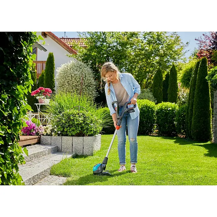 Gardena Power for All 18V Recortabordes de batería EasyCut 23/18V - 18 V, 2 Ah, Iones de litio, 1 batería, Ancho de corte cuchilla: 23 cm - Una mujer usa un cortabordes inalámbrico Gardena para recortar el césped de su jardín. El dispositivo es azul y negro y tiene un mango rojo.