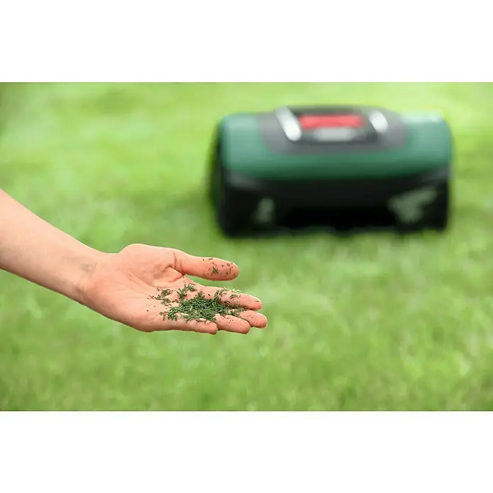 Bosch Mähroboter Indego M 700 - 700 m², Schnittbreite: 19 cm, Induktionsgestützte Navigation, Begrenzungskabel - Ein grüner Rasenmäher-Roboter im Hintergrund mit einer Hand, die Grasschnitt hält. Das Gerät sorgt für einen gepflegten Rasen.