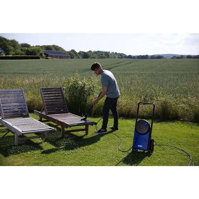 Nilfisk Hidrolimpiadora de alta presión Core 140-6 PowerControl - 1.800 W, Presión máx.: 140 bar, Capacidad de bombeo: 474 l/h - Un hombre limpia una tumbona de jardín de madera al aire libre con una limpiadora de alta presión Nilfisk. Naturaleza verde al fondo.