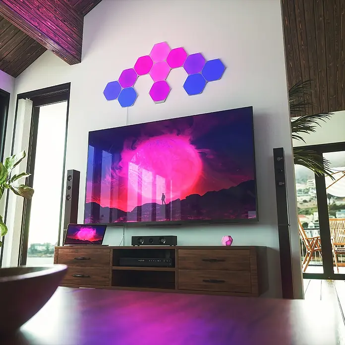 Nanoleaf Erweiterungskit Shapes Hexagons 3 Panels - 3 Stk., Weiß, RGBW, Hexagons, Smart Home-fähig: Ja - Moderne Wohnzimmeransicht mit einem grossen Fernseher, Soundbar und Nanoleaf Lichtpaneelen, die an der Wand befestigt sind, für eine stimmungsvolle Beleuchtung.