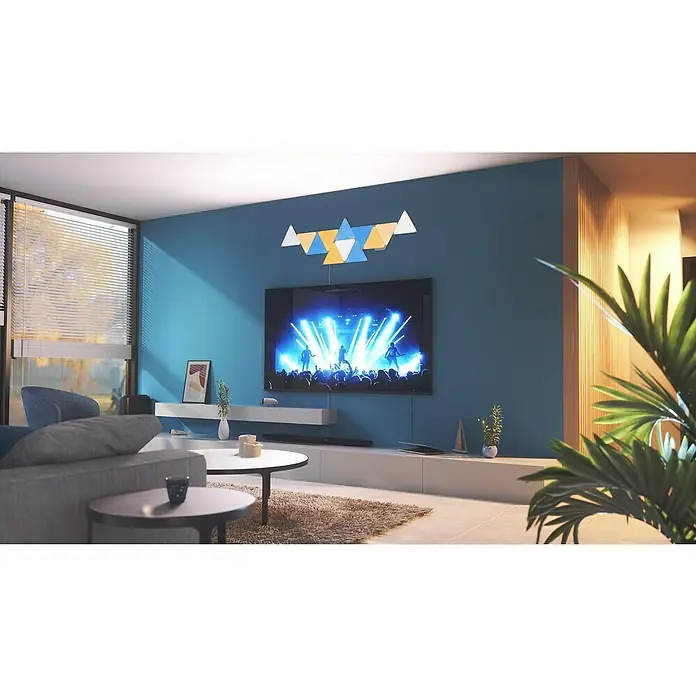 Nanoleaf Erweiterungskit Shapes Triangles 3 Panels - Weiß, RGBW, Triangles, Smart Home-fähig: Ja - Das Bild zeigt ein stilvolles Wohnzimmer mit einem grossen Fernseher und dekorativen, dreieckigen Lichtpaneelen an der Wand.