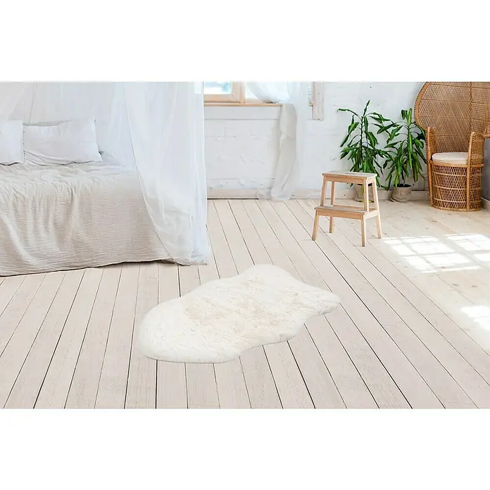 Kunstfell Happy - Ivory, 90 x 60 cm, 100 % Polyester (Flor) - Ein weißer Teppich in Form einer Wolke liegt auf einem hellen Holzboden in einem Schlafzimmer mit einem Bett und Pflanzen im Hintergrund.