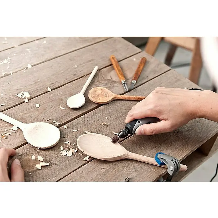 Dremel Multifunktionswerkzeug-Zubehör 681 - 20 -tlg. - Nahaufnahme von Holzlöffeln in verschiedenen Stadien der Fertigstellung auf einem Holztisch, wobei einer mit einem Dremel bearbeitet wird.
