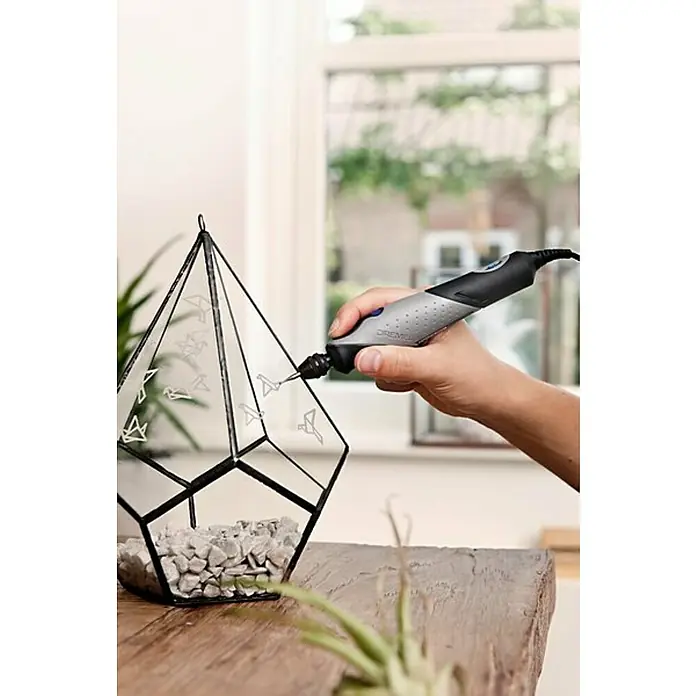 Dremel Glasbearbeitungs-Set 682 - 8 -tlg., Durchmesser Schaft: 3,2 mm - Nahaufnahme einer Hand, die ein Dremel-Rotationswerkzeug verwendet, um ein geometrisches Terrarium mit Origami-Vogelmotiven zu verzieren.