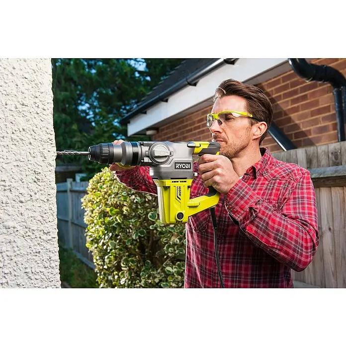 Ryobi Martillo perforador RSDS1050-K - 1.050 W, 3,6 J, Alojamiento SDS-Plus - Un hombre perfora una pared blanca con un martillo perforador Ryobi amarillo, usando gafas de seguridad.