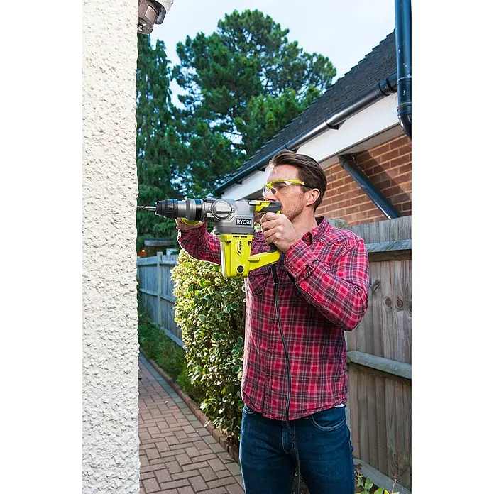 Ryobi Martillo perforador RSDS1050-K - 1.050 W, 3,6 J, Alojamiento SDS-Plus - Un hombre perfora agujeros en una pared exterior con un martillo perforador Ryobi para hacer reparaciones. Lleva gafas protectoras.