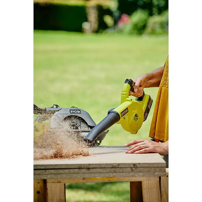 Ryobi ONE+ Akku-Gebläse R18TB-0 - 18 V, Li-Ionen, Ohne Akku - Eine Person benutzt einen gelben Ryobi-Gebläse, um Sägespäne von einem Holztisch zu entfernen. Eine graue Ryobi-Kreissäge steht im Hintergrund.
