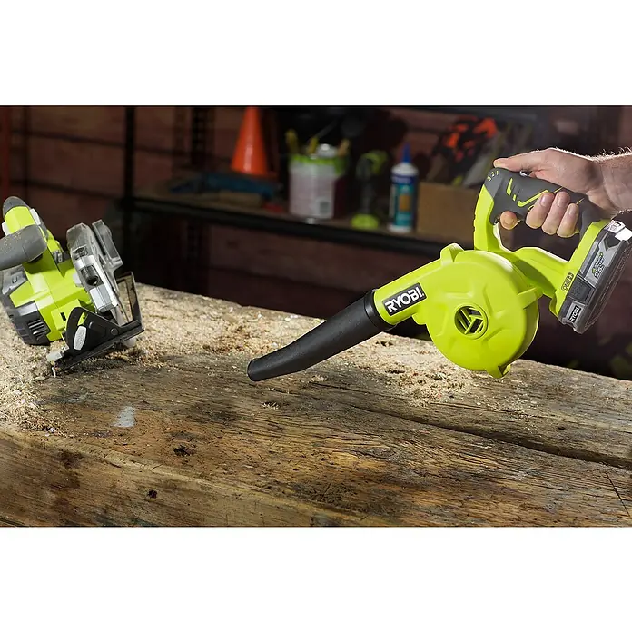 Ryobi ONE+ Akku-Gebläse R18TB-0 - 18 V, Li-Ionen, Ohne Akku - Ryobi Akku-Laubbläser räumt Sägemehl von einem Holztisch, wobei eine Kreissäge im Hintergrund zu sehen ist.