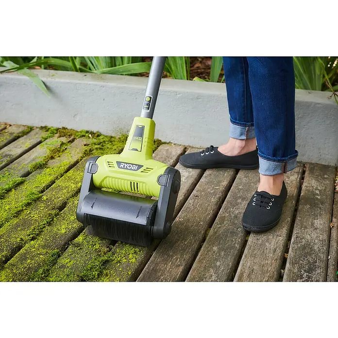 Ryobi ONE+ Oberflächenreiniger RY18PCB-0 - Akkubetrieben, 18 V, Ohne Akku - Nahaufnahme des Ryobi Patios Reinigers, der auf einer Holzterrasse mit Moos verwendet wird, daneben eine Person in Jeans und schwarzen Schuhen.