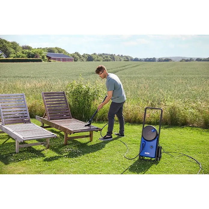 Nilfisk Hochdruckreiniger Core 140-6 PowerControl - 1.800 W, Max. Druck: 140 bar, Fördermenge: 474 l/h - Mann reinigt eine Holzliege mit einem Nilfisk Hochdruckreiniger im Garten, während im Hintergrund ein Feld und ein Haus zu sehen sind.