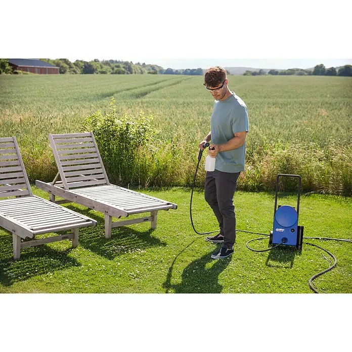 Nilfisk Hochdruckreiniger Core 125-5 Home - 125 bar, 1.400 W, Fördermenge: 438 l/h - Ein Mann benutzt einen Nilfisk Hochdruckreiniger im Freien, um Gartenmöbel zu reinigen. Der Reiniger steht auf dem Rasen neben den Liegestühlen.