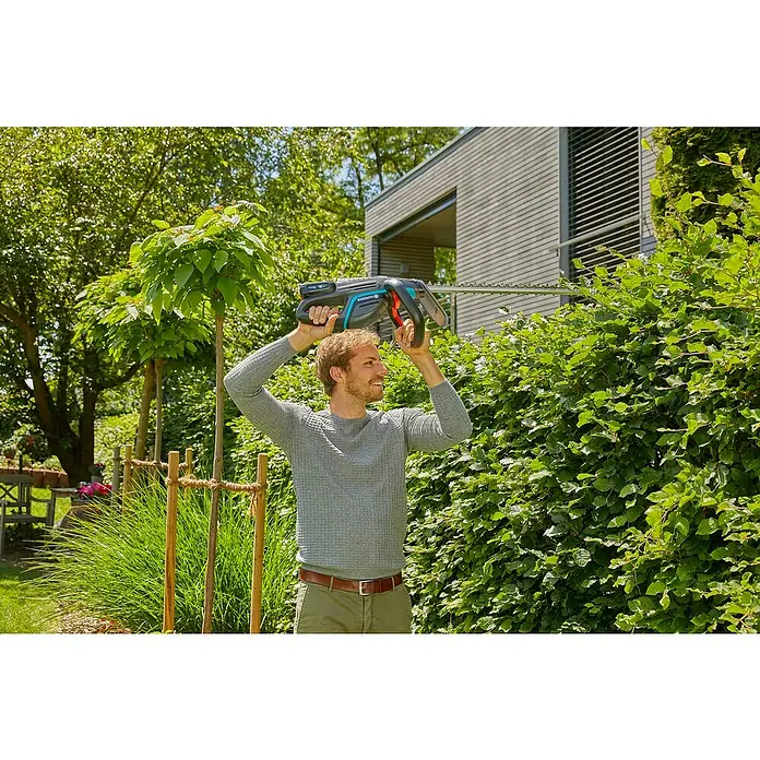Gardena Power for All 18V Akku-Heckenschere ComfortCut 60 - 18 V, Li-Ionen, 1 Akku, Schwertlänge: 60 cm - Ein Mann schneidet mit einer GARDENA Akku-Heckenschere eine Hecke im Garten, während er sie über seinen Kopf hält.