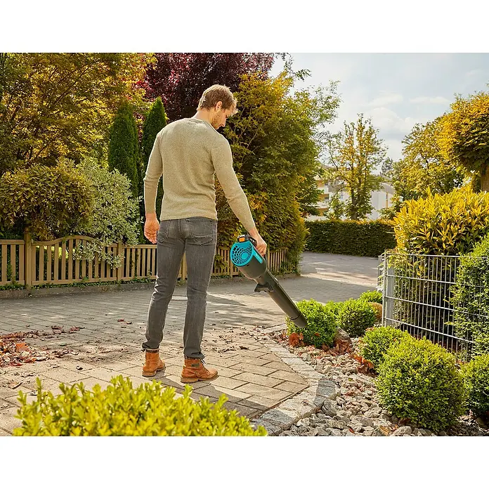 Gardena Power for All 18V Akku-Laubbläser - 18 V, Li-Ionen, 1 Akku, Blasgeschwindigkeit: 100 km/h - Ein Mann benutzt einen Laubbläser der Marke Gardena, um Blätter von einem gepflasterten Weg zu entfernen. Im Hintergrund sind Bäume und Büsche zu sehen.