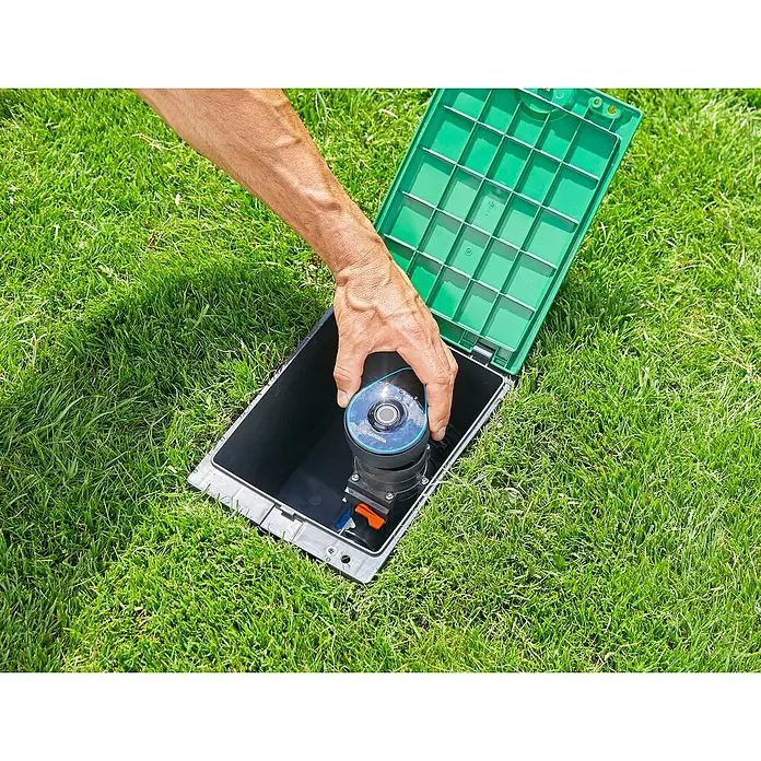 Gardena Sprinklersystem Steuerteil 9 V Bluetooth - Passend für: Gardena Bewässerungsventile 9 V (bis 2020), Gardena Bodenfeuchtesensor - Nahaufnahme einer Person, die einen versenkregner von Gardena in einen unterirdischen Behälter einsetzt, umgeben von grünem Rasen.