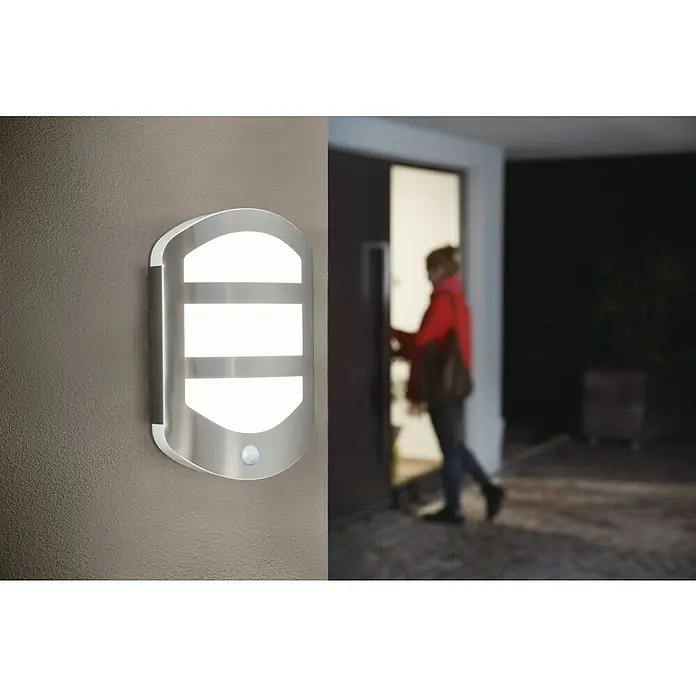 Ledvance LED-Außenwandleuchte Endura - 12,5 W, 180 x 60 x 283 mm, Edelstahl, IP44, Mit Bewegungssensor - Moderne LED-Wandleuchte mit Bewegungssensor außen an einer Hauswand. Das Licht ist eingeschaltet während eine Person die Tür öffnet.