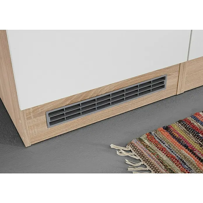 Respekta Küchenleerblock Petunia - Breite: 270 cm, Weiß, Ohne Elektrogeräte - Detailaufnahme eines weißen Schranks mit Holzstruktur und einem grauen Lüftungsgitter an der Unterseite. Davor liegt ein bunter Teppich.