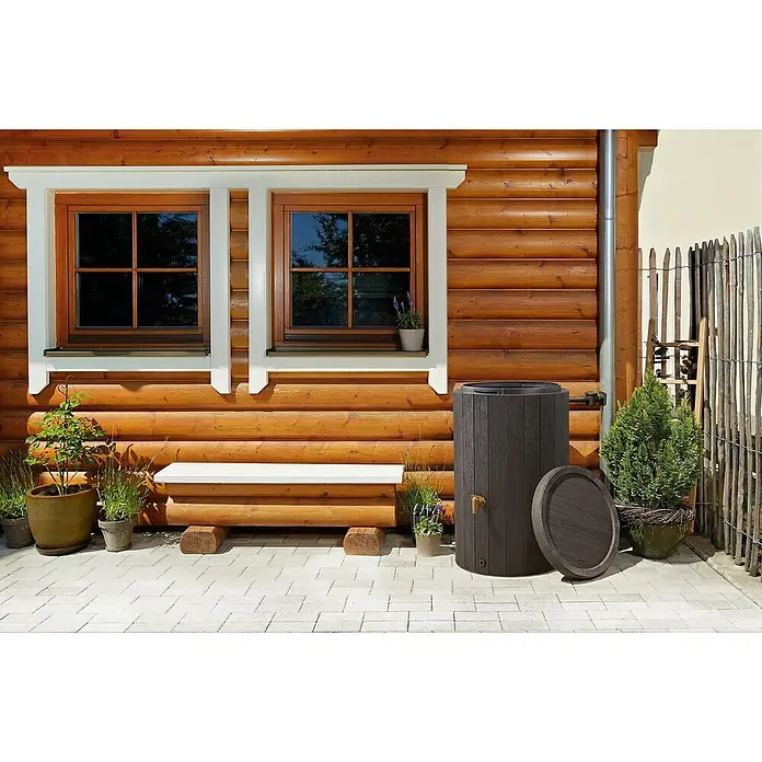 4rain Regenspeicher Madera - 220 l, Dunkelbraun - Eine Regentonne aus dunklem Holzimitat steht neben einem Holzhaus, ideal zur Wassersammlung und Gartenbewässerung.