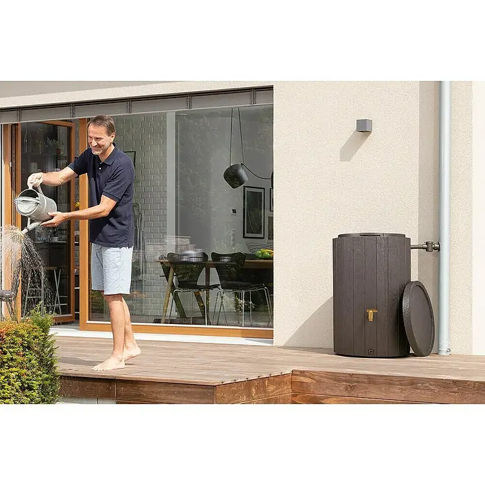 4rain Regenspeicher Madera - 220 l, Dunkelbraun - Ein Mann gießt mit einer Gießkanne aus einem braunen Regenfass, das neben einem Fallrohr auf einer Holzterrasse steht.