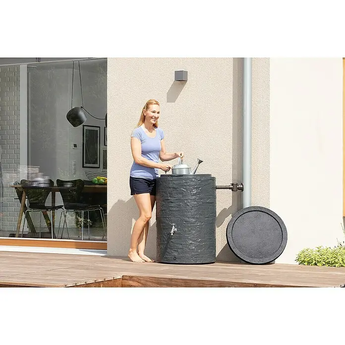 Garantia Classic Line Regenspeicher Muro - 420 l, Graphit - Eine Frau befüllt eine Gießkanne mit einem stilvollen Regenspeicher in Steinoptik, der auf einer Holzterrasse steht.