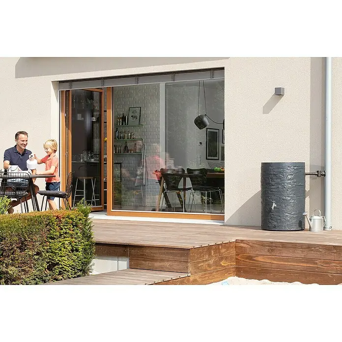 Garantia Classic Line Regenspeicher Muro - 420 l, Graphit - Grauer Regenspeicher mit Wasserhahn und Gießkanne auf einer Holzterrasse vor einem Haus mit Schiebetür.
