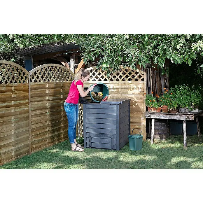 Garantia Komposter Thermo-Wood - 400 l, 72 x 72 x 90 cm, Mit Bodengitter - Kompostierung im Garten: Eine Frau leert einen Eimer in einen dunkelgrauen Kompostbehälter mit Deckel neben einem kleinen grünen Eimer.