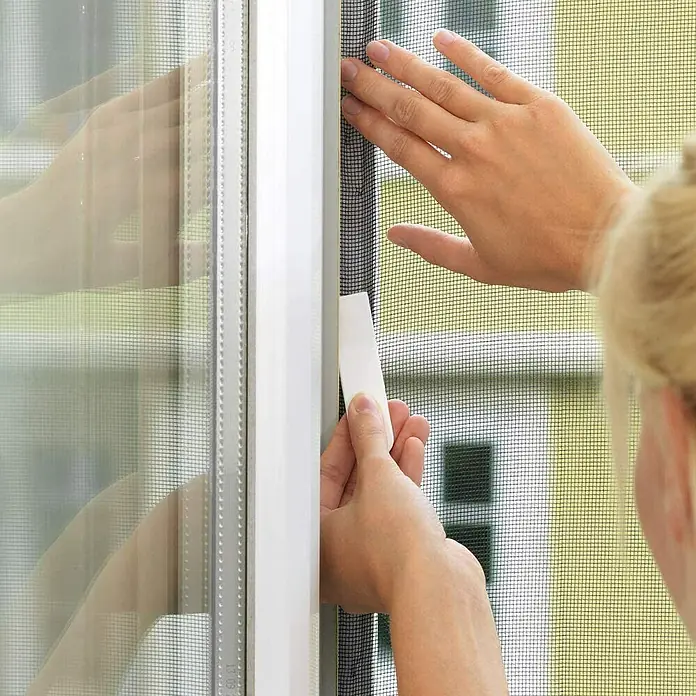 Tesa Insect Stop Cinta para mosquitera Comfort - Largo: 5,6 m, Blanco - Primer plano de una persona que fija una mosquitera a un marco de ventana con una herramienta. La imagen se centra en la instalación de la pantalla.