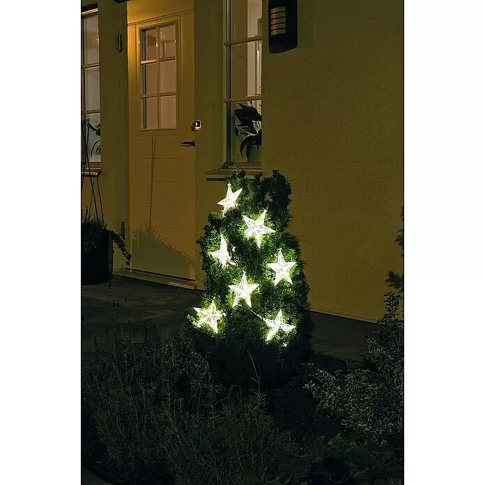 Konstsmide LED-Lichtervorhang - Außen, 5,762 m, Warmweiß, Netzbetrieben - Helle, sternförmige LED-Lichter beleuchten einen immergrünen Strauch im Freien bei Nacht und schaffen eine festliche Atmosphäre.