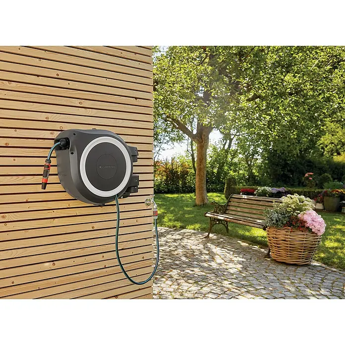 Gardena Wand-Schlauchbox RollUp M/L - Schlauchlänge: 25 m - Das Bild zeigt eine automatische Gardena-Schlauchbox an einer Holzwand im Garten, mit Bank, Blumen und Bäumen im Hintergrund.