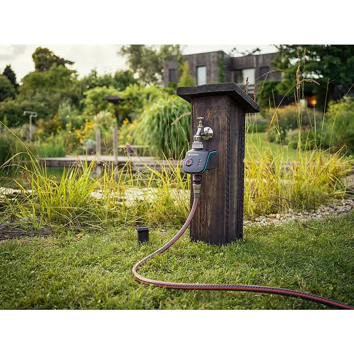 Gardena Smart system Bodenfeuchtesensor Control Set - Passend für: Verbindung von elektrischen Geräten aus dem Gardena smart system - Nahaufnahme eines Gardena Smart Water Control-Systems an einem hölzernen Wasserhahn im Garten, umgeben von üppigem Grün.