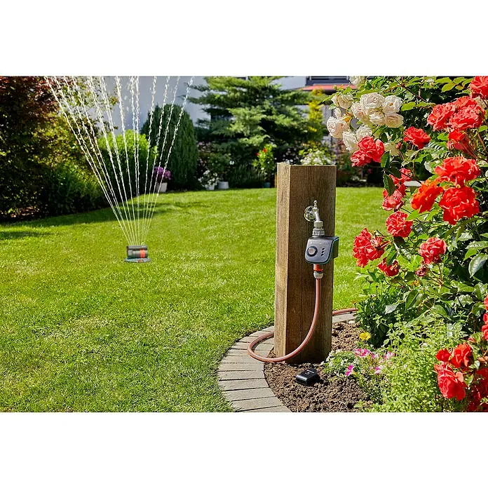 Gardena Smart system Bodenfeuchtesensor Control Set - Passend für: Verbindung von elektrischen Geräten aus dem Gardena smart system - Eine Gardena Bewässerungssteuerung ist an einem Holzpfosten im Garten befestigt, während ein Rasensprenger im Hintergrund arbeitet.