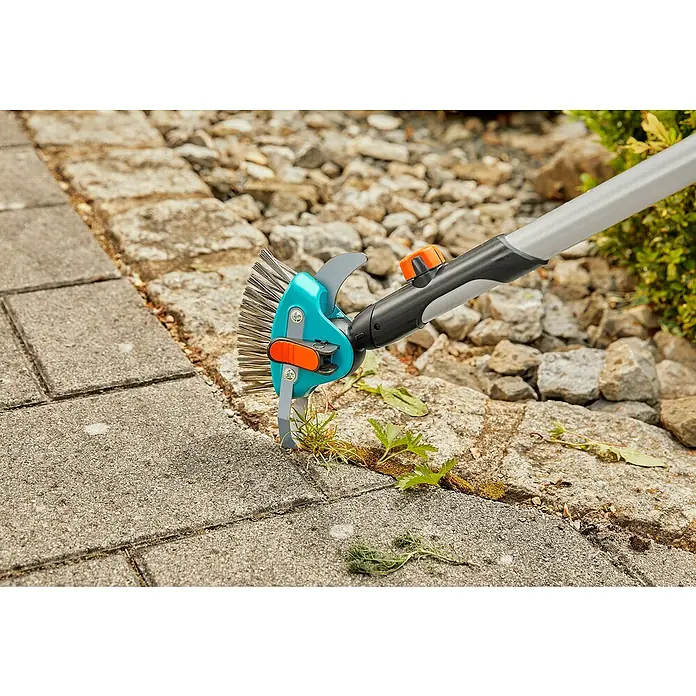 Gardena Combisystem Fugenkratzer 3in1 - Länge: 23,5 cm, Material Gerätekopf: Edelstahl - Das Bild zeigt einen Gardena Fugenkratzer im Einsatz, um Unkraut zwischen Pflastersteinen zu entfernen. Das Werkzeug ist blau und grau mit orangefarbenen Akzenten.