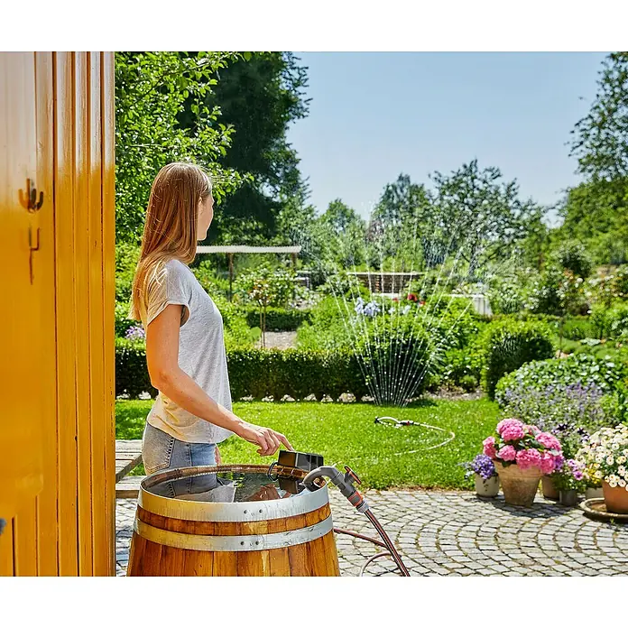 Gardena Power for All 18V Akku-Regenfasspumpe - 18 V, 1 Akku, Max. Fördermenge: 2.000 l/h - Frau nutzt ein Gardena Bewässerungssystem mit einem Holzfass für eine umweltfreundliche Gartenbewässerung.