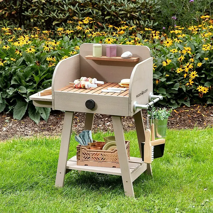Muddy Buddy Spielgrill BBQ Chef - 68 x 37 x 79 cm, Warmgrau - Ein detailreicher Spielzeuggrill von Muddy Buddy mit Zubehör vor blühenden Blumen. Perfekt für fantasievolle Outdoor-Rollenspiele.