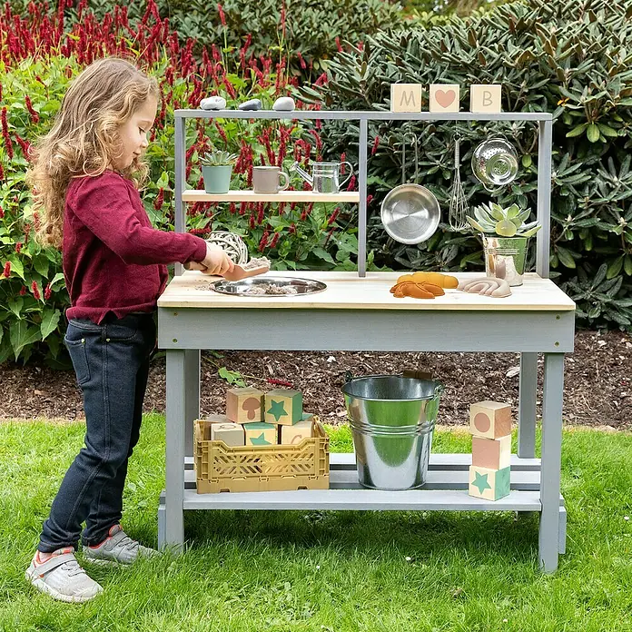 Muddy Buddy Matschküche Mud Starter - 80 x 40 x 90 cm, Holz, Natur/Wolkengrau - Ein Kind spielt mit einer Outdoor-Spielküche, die mit Töpfen, Pflanzen und Spielzeugblöcken ausgestattet ist.