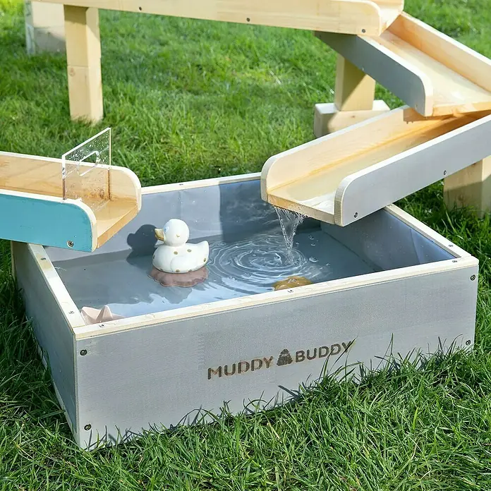 Muddy Buddy Wasserbahn Aqua Traveller - Holz, Natur/Wolkengrau - Nahaufnahme eines Muddy Buddy-Wasserspieltisches mit einem Spielzeugenten im Wasser, auf einer grünen Wiese.